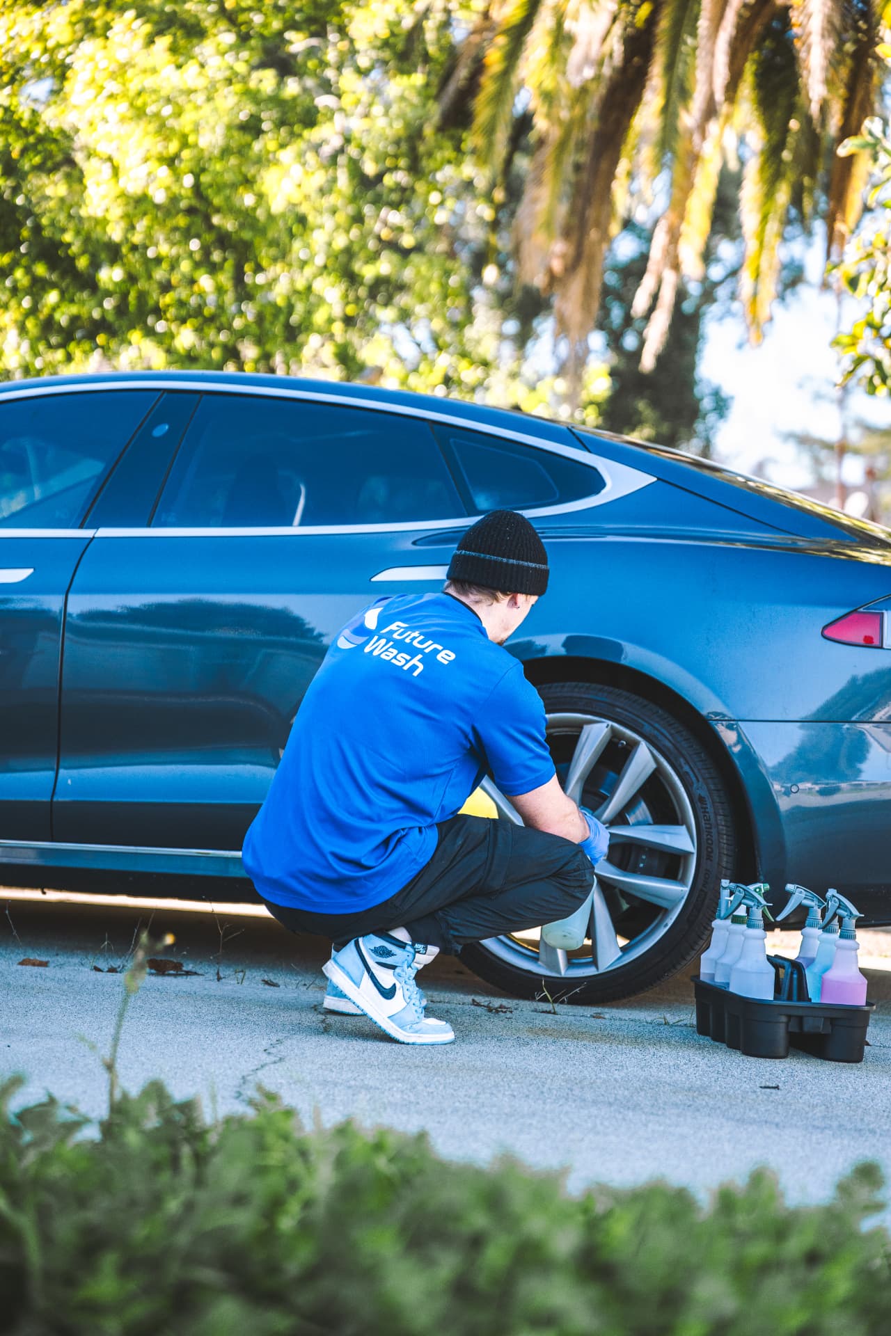 Future Wash technician crouching to clean the wheel of a dark Tesla in a driveway